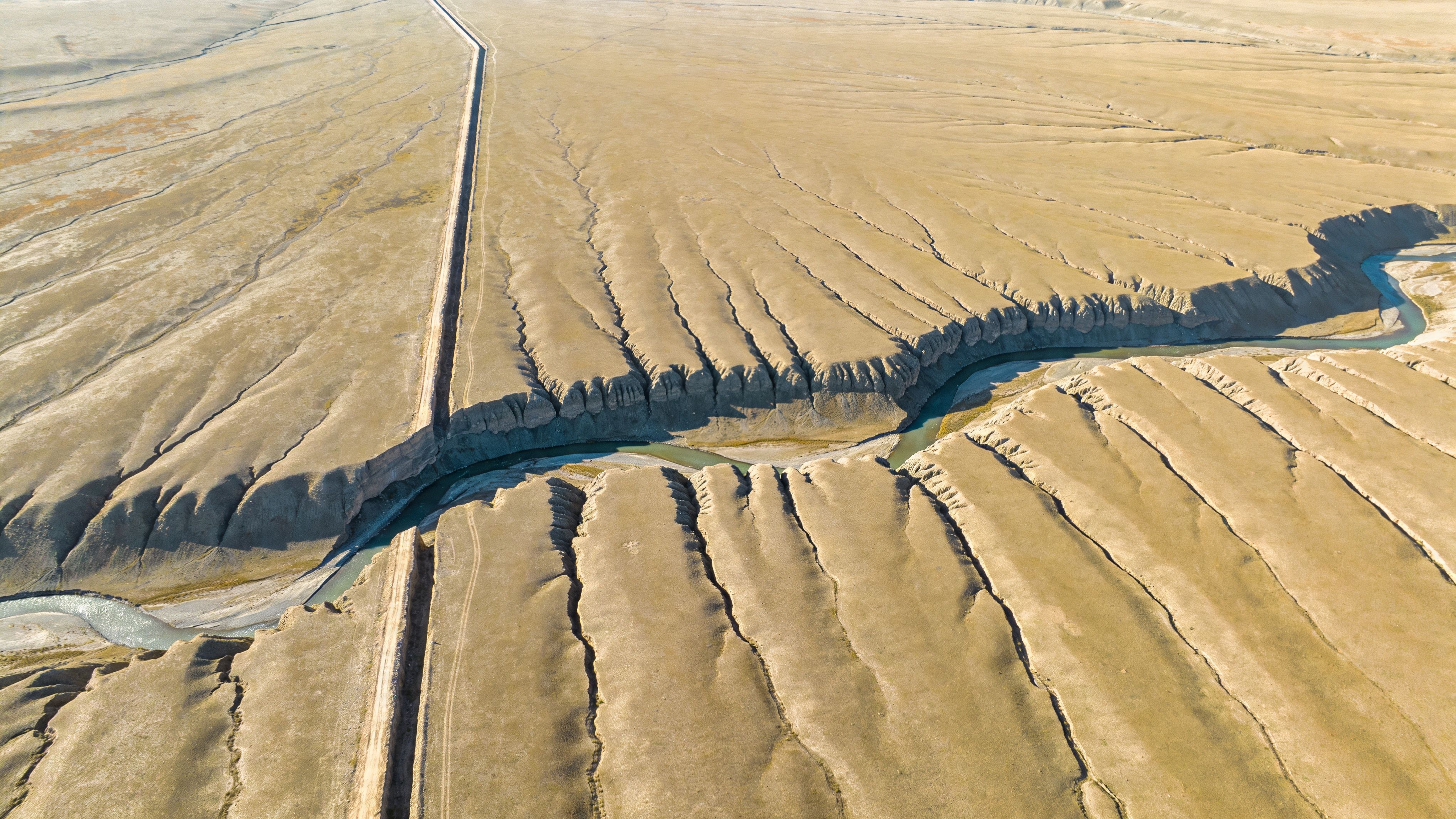 An aerial view of a river cutting through arid, cracked desert landscape near a long road.