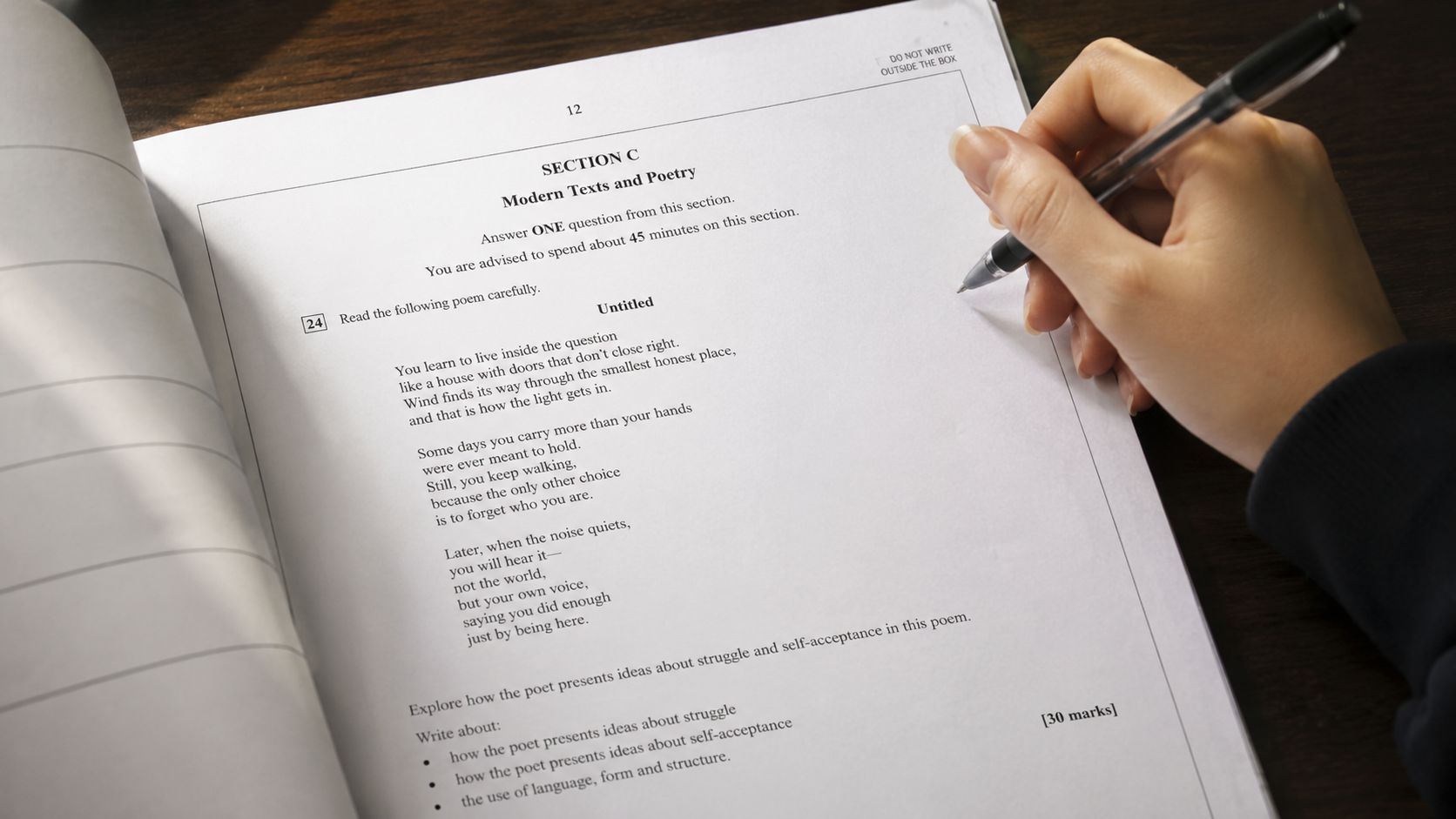 A hand writes on an exam paper featuring a poem about struggle and self-acceptance on a desk.
